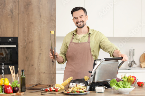 Young happy man cooking fresh corn cobs on modern electric grill in kitchen