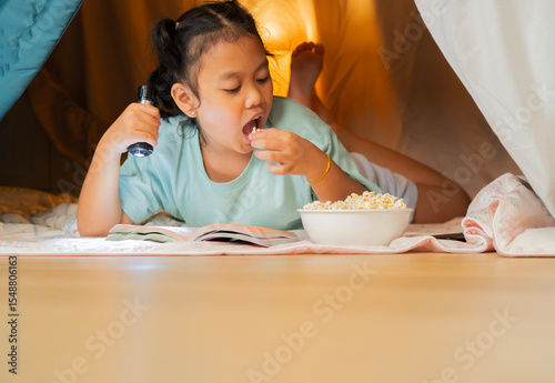 Little girl eating popcorn while enjoying reading a book on the floor under a table.