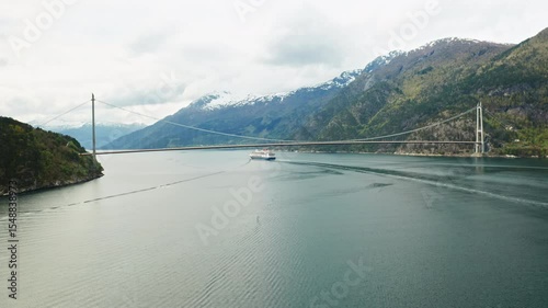 Hardanger Bridge stretches over fjord and white cruise ship sails under bridge to distance. Mountains and overcast sky highlight landscape of Norway
