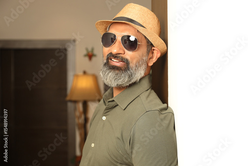 Mid shot of happy middle age handsome man with hat, sunglasses posing at home while looking at the camera Indian stylish beard man posing model facial expressions and body language 