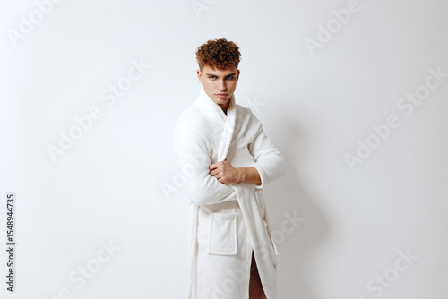 Confident young man posing in a white bathrobe against a plain light background, showcasing self-assurance and style. Fashion and wellness concept.