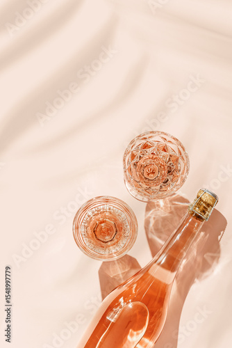 Glasses of rose wine on pink background with palm leaf shadows from sunlight. Minimal Top view Vintage wineglasses and bottle, sparkling alcohol drink above view, caustics from hard light, copyspace