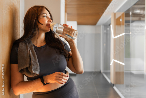 Sporty woman hydrating with water after workout, leaning on wall in modern gym interior