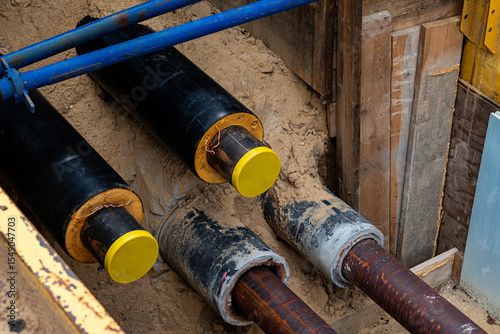 Construction workers are installing utility pipes in a deep trench filled with sand. The pipes are secured with wooden supports, indicating a significant underground infrastructure upgrade.