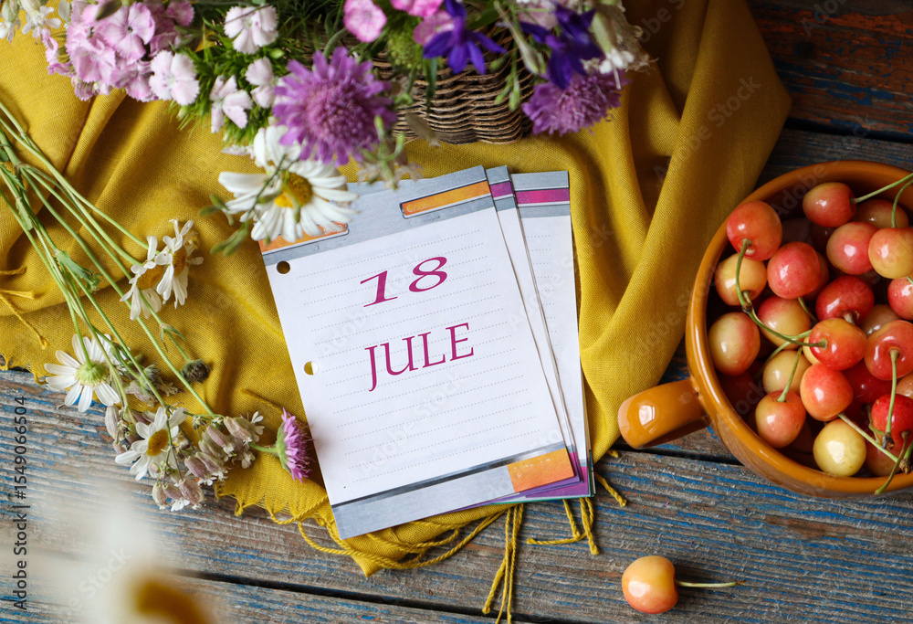 Naklejka premium Calendar for July 18: flip calendar with numbers 18, name of the month July in English on a yellow napkin with wild flowers and ripe cherries