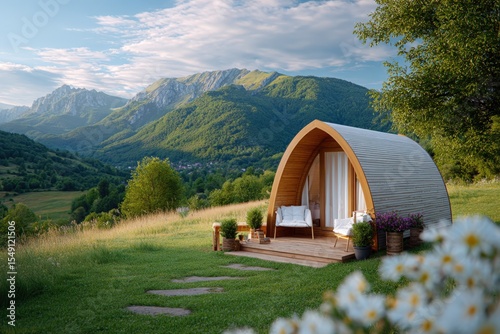 Scenic landscape with green mountains lush grass trees and a modern tiny wooden cabin with a curved roof and outdoor patio area in a peaceful countryside setting