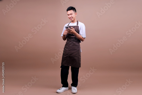 Full Length Of Smiling Young Asian Guy In Brown Apron Using Smartphone While Standing