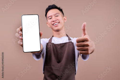 Smiling Young Man In Brown Apron Showing Blank Smartphone And Giving Thumbs Up Over Beige Background