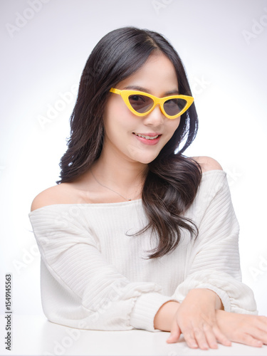 Asian young beautiful attractive cute happy woman long black hair poses with smile cheerful expression, wearing white sweater yellow sunglasses fashion, sitting white table against light background.