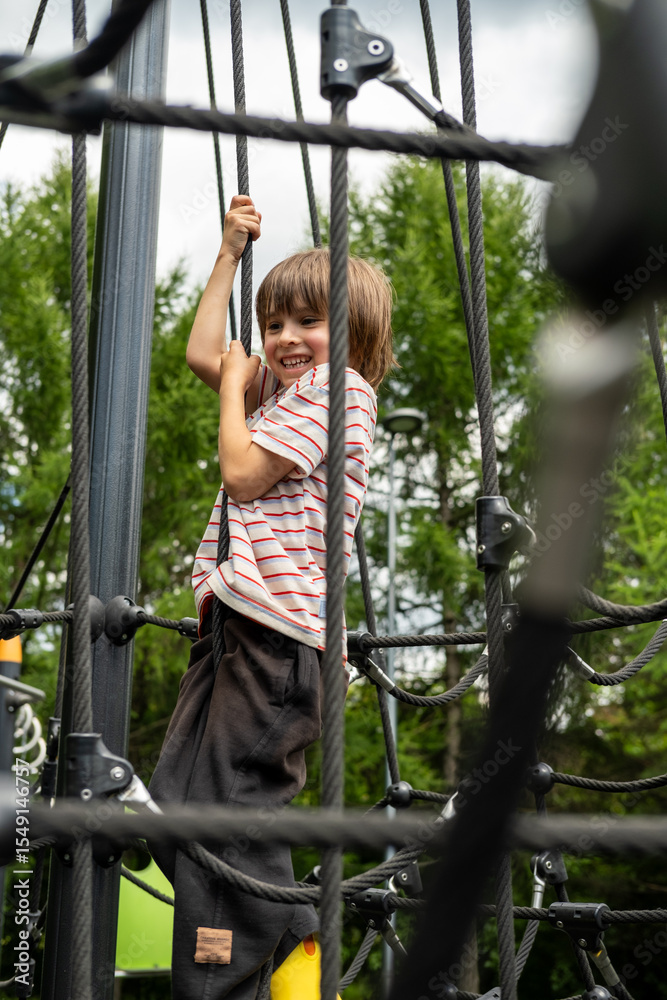 Obraz premium A child boy is climbing up an alpine grid in a park on a playground on a hot summer day. Kids playground in a public park, entertainment and recreation for children, mountaineering training.