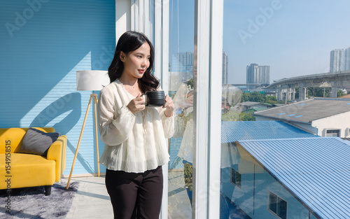 Asian young beautiful happy woman wears casual clothes standing holding drinking coffee cup or hot beverage look out glass window in light blue living room at home relax morning coffee warming hands.