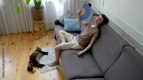 Exhausted woman fanning herself on sofa while cat lying nearby in heat distress. Indoor heat suffering, pet owner and animal under stress, summer heat without AC, discomfort, dehydration at home