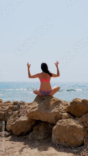 Young woman in swimsuit meditates, practicing yoga, while sitting on stone on beach by the sea. body care and meditation concept