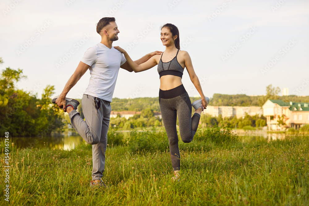 Smiling young man and woman doing leg stretching exercises together on grass. Athletic couple warming up before workout session, supporting each other and enjoying healthy active lifestyle outdoors. Smiling young man and woman doing leg stretching exercises together on grass. Athletic couple warming up before workout session, supporting each other and enjoying healthy active lifestyle outdoors.