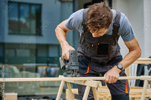Foto Man cutting wooden plank with electric jigsaw at home backyard