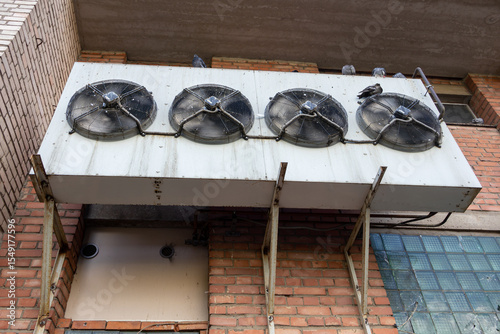 Industrial air conditioner with four fans are mounted on a building. The fans are black and the building is brick. Pigeons are sitting on the air conditioner