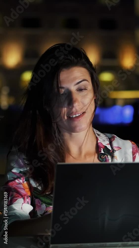 Beautiful young woman uses laptop and listens to music, singing along, dancing with delight. She sitting at open terrace or balcony, against background of night city bright lights. warm summer night.