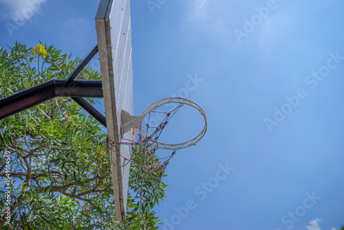 An old outdoor basketball hoop with a weathered wooden backboard captured from below under a bright blue sky. The frayed net and rustic look evoke nostalgia and street basketball vibes.