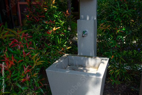 An outdoor sink with a metal faucet surrounded by green and red ornamental plants. The modern stone structure is situated in a shaded garden area, evoking cleanliness and nature.