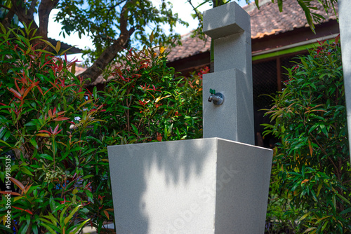 An outdoor sink with a metal faucet surrounded by green and red ornamental plants. The modern stone structure is situated in a shaded garden area, evoking cleanliness and nature.