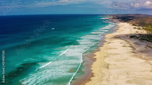 Aerial view of a pristine coastline with turquoise waves gently washing onto the sandy shore, under a bright blue sky, evoking summer relaxation and seaside adventure