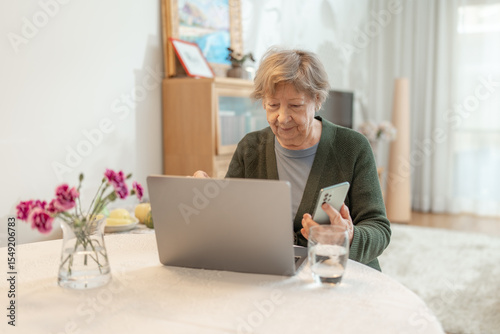 Senior woman using laptop at home while holding a smartphone. Concept of technology, online shopping, digital literacy for seniors and active aging. Technology usage among seniors.