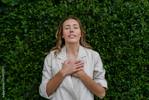 Relaxed woman standing against a green hedge with a serene expression