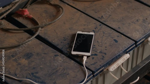 Mobile phone is lying on dirty and dusty surface and is being charged via wire. Close-up. Adapter is plugged into socket and converts electricity for phone's battery