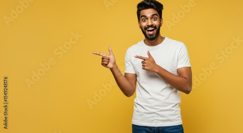Excited Man Pointing Sideways - A young man with a joyful expression points to the side against a vibrant yellow background. He's wearing a plain white t-shirt and jeans