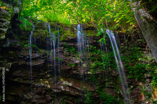 A bluff overhanging the Keown Falls Loop Trail in Chattahoochee National Forest, with small streams of water falling over the edge and sunlit trees in the background