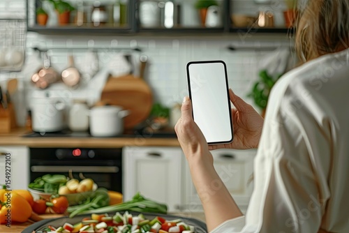 Woman is Holding Smartphone with Blank Screen for Your Design During Cooking in Modern Kitchen