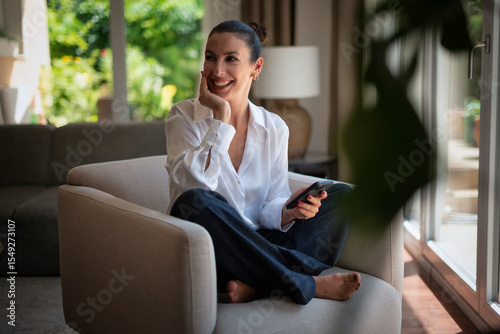 Smiling brunette haired woman relaxing in an armchair in her luxury home and using phone