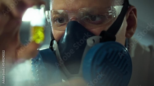 A scientist investigates a bright yellow liquid in a lab. Equipped with a respirator and goggles, he focuses intently on the experiment