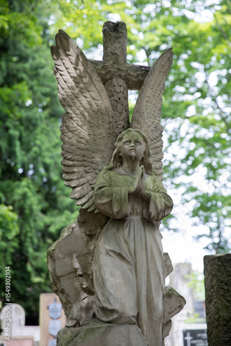 Weathered stone angel statue with wings and a wreath in an old cemetery. Surrounded by green trees and gravestones. Peaceful, somber atmosphere with historic and spiritual symbolism.