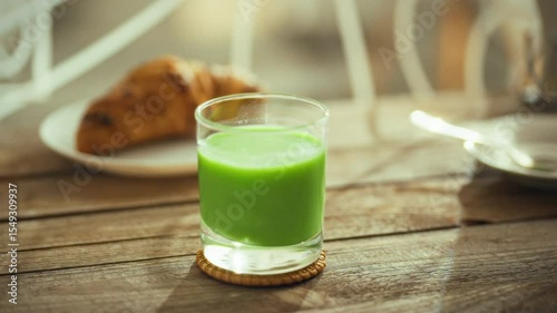 A chilled green beverage is placed on a coasters next to a golden croissant, creating a cozy setup on a wooden table. The atmosphere reflects a relaxed, casual dining experience