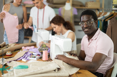Positive middle-aged man sewing while others working with sewing-machine and making details in dressmaking courses