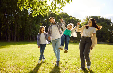 Happy family walking and playing together outdoors in summer green park, parents swinging small child, playfully kids, friendly smiling enjoying time together on sunny day weekend fun, happiness 
