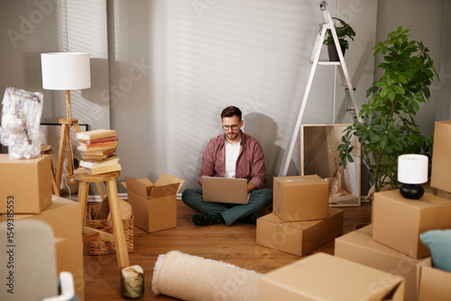 Obraz na płótnie A person sits on the floor amidst unpacked cardboard boxes, using a laptop to organize tasks after moving to their new residence