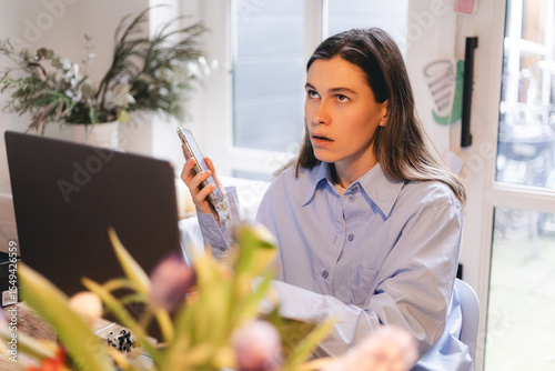 Woman sitting at home desk, talking by mobile phone, working near laptop, she listen voice message and rolling her eyes. Angry annoyed girl.