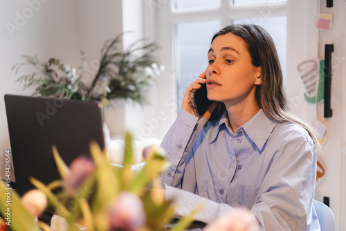 Foto Serious businesswoman talking on mobile phone at home workplace, working at computer