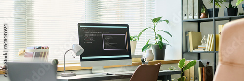 Modern workspace featuring large desktop monitor displaying neural network code, desk lamp, potted plants, bookshelves in background, sunlight streaming through window blinds