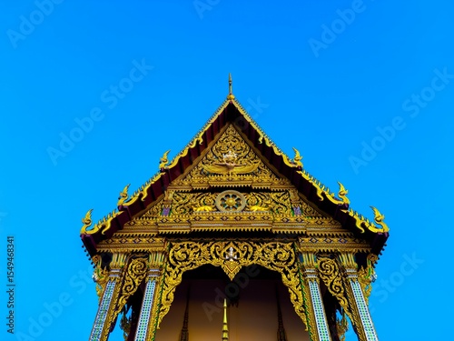 A low-angle, head-on view captures the resplendent golden gable of a traditional Thai temple, set against an expanse of clear, azure blue sky. The intricate details of the carved and gilded