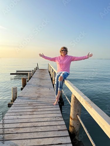 young woman with arms outstretched sitting on wooden pier in sunset, freedom and travel concept