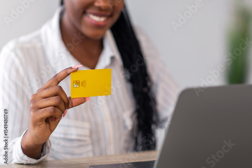 Credit card in cheerful black lady hand, pretty african american young woman in official outfit using laptop at home, having remote job, freelancer using online banking or shopping online, empty space