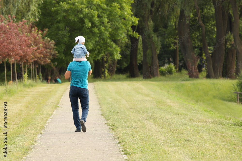 Fototapeta premium A father and his little child are walking along the alley of a summer park. A man and a girl are walking in nature. Relaxation in nature for the harmonious development of a child. Relationship between
