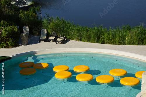 Beneath a radiant sky, a large public swimming pool stretches out, lined with relaxed swimmers and colorful lounge chairs. The sun sparkles on the water’s surface, creating shimmering reflections that