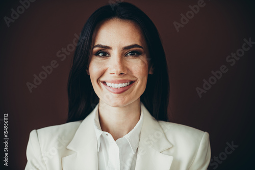 Elegant professional young woman in formal white blazer smiling confidently against brown background symbolizing career success