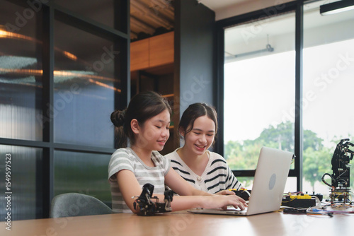 Two young Diverse girls learning to program robot arms on laptop in a stem education class, smiling and studying coding skills in a modern indoor technology