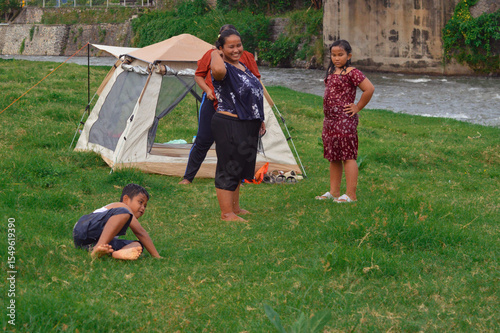 A playful moment captured as the young boy falls forward on the grass, and the nearby group standing by the tent bursts into laughter at his act.