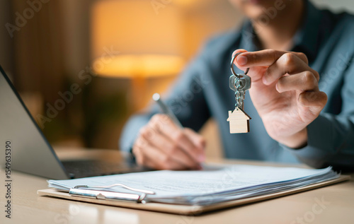 Person holds a house-shaped keychain while signing property agreement next to a miniature house model, laptop and clipboard on desk.